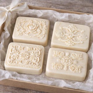 May include: Four ivory-coloured, square soaps with floral and ornate designs are presented in a brown box lined with tissue paper. A cream-coloured ribbon is tied in a bow on the box. The soaps have embossed patterns of roses and other decorative elements.