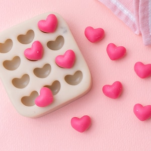 May include: A silicone mould with 14 heart-shaped cavities, filled with pink heart-shaped sweets. The mould is on a pink surface, and there are several pink heart-shaped sweets scattered around it.