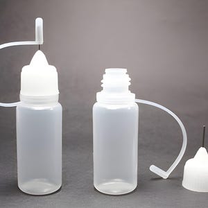 May include: Three clear plastic bottles with white caps and dispensing tips. One bottle has a cap with a needle tip, another has a cap with a needle tip attached, and the third bottle is uncapped. The bottles are on a grey background.