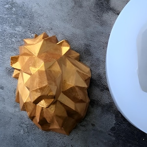 May include: A gold geometric lion head sculpture with a detailed, faceted design. The sculpture is made of a smooth, shiny material.