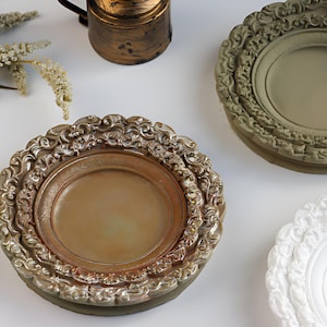May include: A collection of decorative plates in gold, green, and white. The plates feature intricate, sculpted edges and come in different sizes. A small, antique-style metal container is also visible.