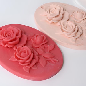 May include: Two oval-shaped silicone molds for making decorative rose-shaped soap or candles. The mold on the left is a bright pink color and the mold on the right is a light pink color.
