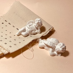 May include: Two white cherub figurines with detailed wings and curly hair positioned on a piece of light beige fabric. The fabric features a printed calendar grid with black text showing days of the week and numerical dates.