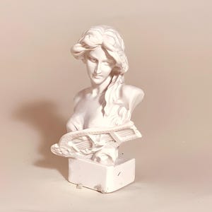 May include: A white bust sculpture of a woman with flowing hair, looking down. The sculpture includes a lyre and is mounted on a rectangular base. The bust is a classical art piece.