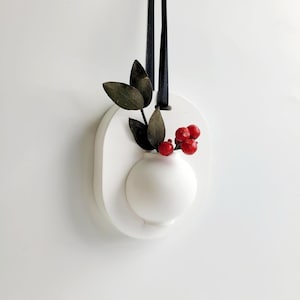 May include: A white, oval wall decoration featuring a small, round vase. The vase contains dark green leaves and red berries. A black ribbon is attached for hanging. The design is minimalist and decorative.