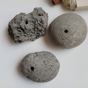 May include: Three gray stones with natural textures and porous surfaces, each featuring a single drilled circular hole. The stones vary in shape, with one being jagged and irregular, while the other two are rounded and smooth in appearance.