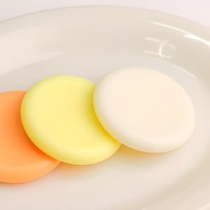 May include: Three round soaps on a white plate. The soaps are yellow, orange, and white.