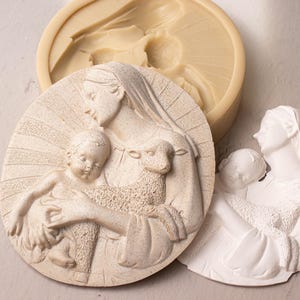 May include: A white plaster mold of a religious scene depicting a mother holding a child and a lamb. The mold is on a light gray surface.