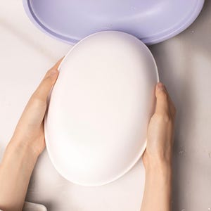 May include: A person is holding a white oval-shaped plate. The plate is smooth and has a slight curve to it.