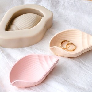 May include: Three seashell-shaped trinket dishes, one beige, one pink, and one white. The beige dish has two gold rings inside.