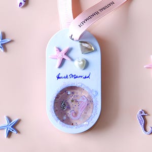 May include: A light blue oval-shaped ornament with a pink starfish, a pearl heart, a clear rhinestone, and a clear dome filled with glitter and a pink seahorse. The ornament has the words "Just Married" written on it and is attached to a pink ribbon with the words "Natural Handmade".