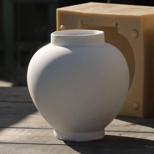 May include: A white ceramic vase with a rounded shape. The vase is sitting on a wooden surface.