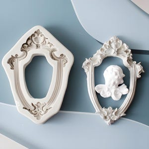 May include: Two white silicone moulds for baking. The moulds are shaped like ornate frames with intricate details. One mould features a cherub angel head in the centre.
