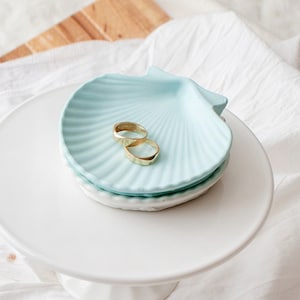 May include: A light blue ceramic seashell dish with two gold rings inside. The dish is on a white cake stand.
