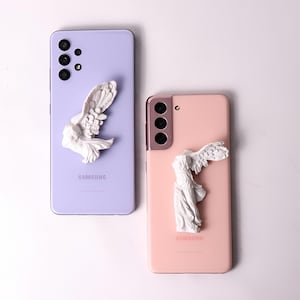 May include: Two Samsung Galaxy smartphones with different colored cases. The phone on the left has a lavender case with a white angel figurine on top. The phone on the right has a pink case with a white angel figurine on top. Both phones have the Samsung logo on the back.