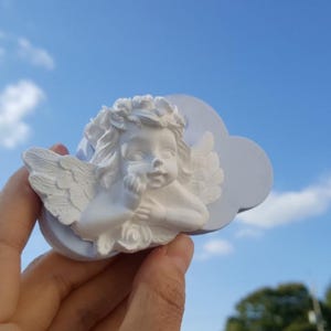 May include: A white silicone mould featuring a detailed relief of a cherub with wings and curly hair, set against a cloud-shaped background. The mould is designed for casting decorative figures and has a smooth, matte finish on its surface.