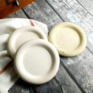 May include: A collection of ceramic coasters in various shades of cream and beige. Two coasters are a solid cream color, while the third has a speckled beige appearance. They are arranged on a white cloth with a red stripe.