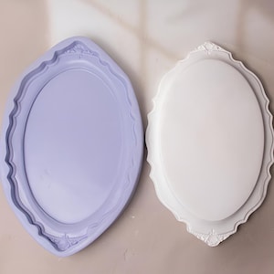 May include: Two oval-shaped molds, one lavender and one white, with ornate, scalloped edges. The lavender mold has a detailed floral design on the frame. The white mold has a smooth surface. Both are likely for crafting or decorative purposes.