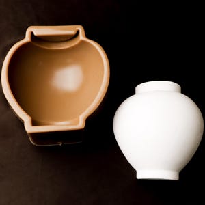 May include: Two ceramic vases. One is a light brown, open-topped vessel with a handle-like design. The other is a white, rounded vase with a short neck and a flat base. Both are on a dark brown and black background.