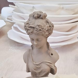 May include: A ceramic bust of a woman with detailed hair and a draped garment. The bust is painted in a light brown colour.