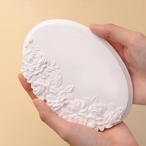 以下が含まれることがあります： 縁に浮き彫りの花柄が施された白い楕円形のセラミック皿。皿は両手で持たれています。