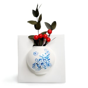 May include: A white ceramic wall-mounted vase with a square backplate and a rounded front featuring blue floral patterns. The vase contains a decorative stem with dark green leaves and several small, round, bright red berries.