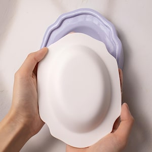 May include: A white ceramic bowl with a scalloped edge is being held by a person's hands. The bowl is being placed on top of a light purple ceramic bowl with a scalloped edge.