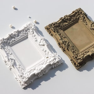 Peut inclure: Deux cadres photo rectangulaires avec des détails floraux et des volutes en relief. L'un des cadres est blanc uni, tandis que l'autre est d'une teinte brune terreuse et mate. Plusieurs petites perles blanches rondes sont éparpillées.