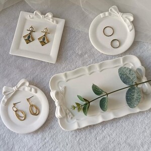 May include: A collection of four white ceramic trays featuring sculpted bow accents. The set includes one square tray, two circular trays, and one rectangular tray with scalloped edges, displayed with gold-toned earrings and rings.