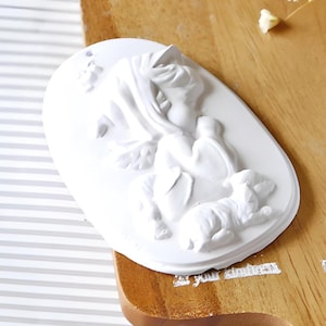 May include: White ceramic mold with a raised design of a woman holding a small dog. The mold is oval shaped and has a smooth surface.