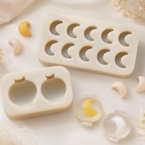 May include: Two off-white silicone molds, one rectangular with ten crescent moon cavities and one smaller with two circular jar-shaped cavities. Several small, solid crescent moon shapes in yellow and cream colors are scattered nearby.