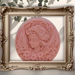 May include: A pink, circular soap with a carved angel design, set within an ornate gold-coloured frame. The angel has closed eyes, flowing hair, and is surrounded by wings and flowers. The frame has decorative detailing.