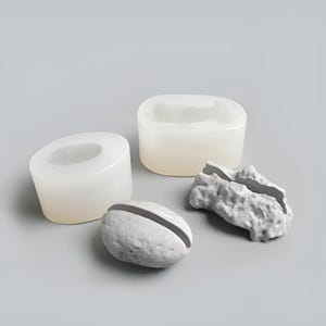 May include: Two translucent white silicone molds and two gray, textured, rock-like objects with a central slit. The molds are cylindrical with hollow centers, while the gray objects feature irregular, rough surfaces and a deep groove running across.
