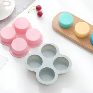 May include: Two silicone moulds for making round treats. One mould is pink and has four cavities. The other mould is grey and has four cavities. There are three round treats on a wooden board. The treats are blue, yellow, and light blue.