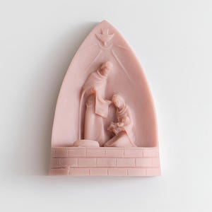 May include: A pink ceramic nativity scene with a dove above the figures of Mary, Joseph, and baby Jesus. The scene is set in a brick stable.