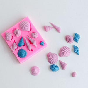 May include: A square pink silicone mold featuring various seashell-shaped cavities, accompanied by several loose, hardened seashell-shaped castings in shades of light pink and bright blue, scattered across the surface next to the mold.