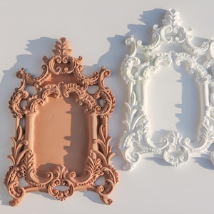 May include: Two ornate picture frames, one in a light brown colour and the other in white. Both frames have a detailed, intricate design with a baroque style.