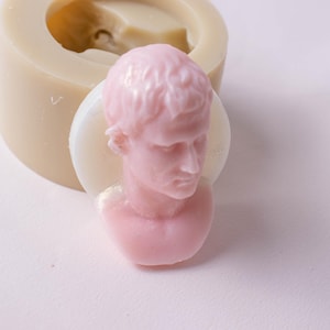 May include: A light pink soap bust of a classical figure, set on a white disc. A beige silicone mould is in the background. The soap has detailed facial features and hair. The image is well-lit, highlighting the soap's texture and form.