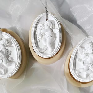 May include: Three white ceramic oval-shaped air fresheners with a raised design of a cherub angel. Each cherub is holding a different object, one is holding a flower, one is holding a ribbon, and one is holding a heart. The air fresheners are hanging from a string.