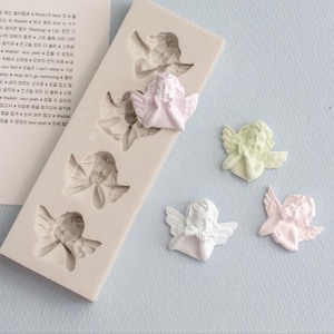 May include: A rectangular silicone mould featuring four recessed angel cavities, accompanied by three small, individual angel figures in pastel pink, pale green, and white. The figures depict cherubs with folded wings and hands held in a prayer position.