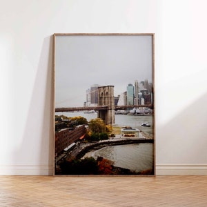 Puede incluir: Una fotografía enmarcada del puente de Brooklyn en la ciudad de Nueva York. El puente se ve desde la distancia, con el horizonte de la ciudad en el fondo. La foto fue tomada en un día nublado, y el agua es de color gris oscuro.