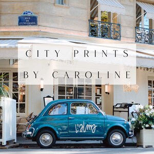 May include: A teal blue Fiat car parked in front of a building with a white awning that says "CITY PRINTS BY CAROLINE". The car has a white sticker on the side that says "Lalma".