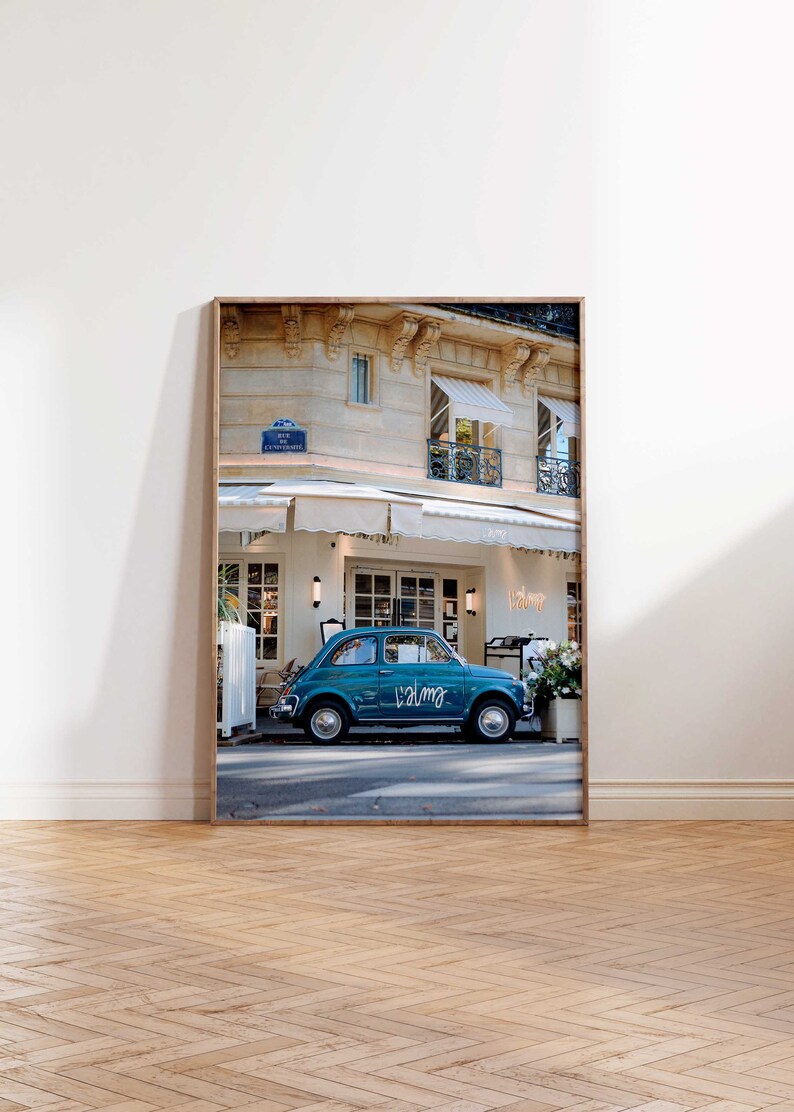 May include: A teal blue vintage car parked in front of a cafe in Paris, France. The car has the word "Calme" written on the side. The cafe has a white awning and a sign that says "Valmy".