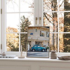 May include: A framed print of a teal blue vintage car parked in front of a building with a sign that reads "Volare". The print is on a white windowsill with a candle, a small vase, a book, and a wooden bowl.