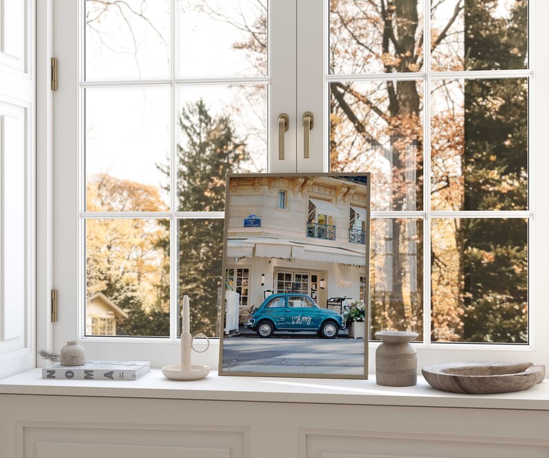May include: A framed print of a teal blue vintage car parked in front of a building with a sign that reads "Volare". The print is on a white windowsill with a candle, a small vase, a book, and a wooden bowl.