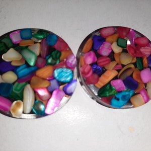 May include: Two round, clear resin coasters with a colorful mosaic of iridescent shell pieces embedded inside. The coasters are approximately 2 inches in diameter.
