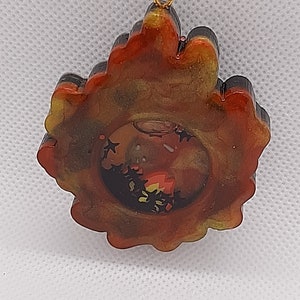 May include: A gold-toned pendant with a black and red resin design. The resin has a swirling pattern and a clear center with glitter. The pendant is attached to a gold chain.