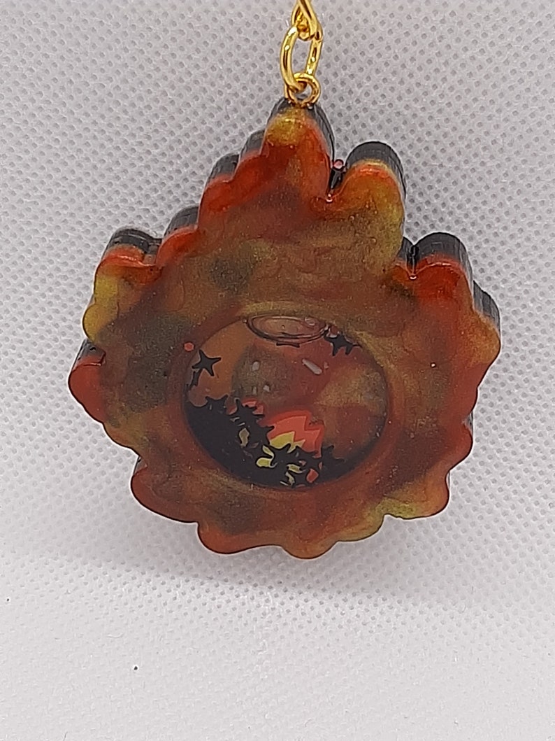 May include: A gold-toned pendant with a black and red resin design. The resin has a swirling pattern and a clear center with glitter. The pendant is attached to a gold chain.