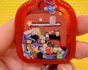 May include: Red resin keychain shaped like a backpack filled with colorful school supplies, including pencils, crayons, erasers, and a star. The keychain has a clear plastic badge holder and a metal clip.