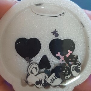 Könnte beinhalten: Ein weißer, glitzernder Schädel-Shaker mit schwarzen Herzaugen und einer schwarzen Nase. Im Inneren befinden sich verschiedene Halloween-Konfetti-Stücke, darunter Knochen, Kürbisse und ein Grabstein mit "RIP". Der Shaker hat eine klare, blasenartige Oberseite.